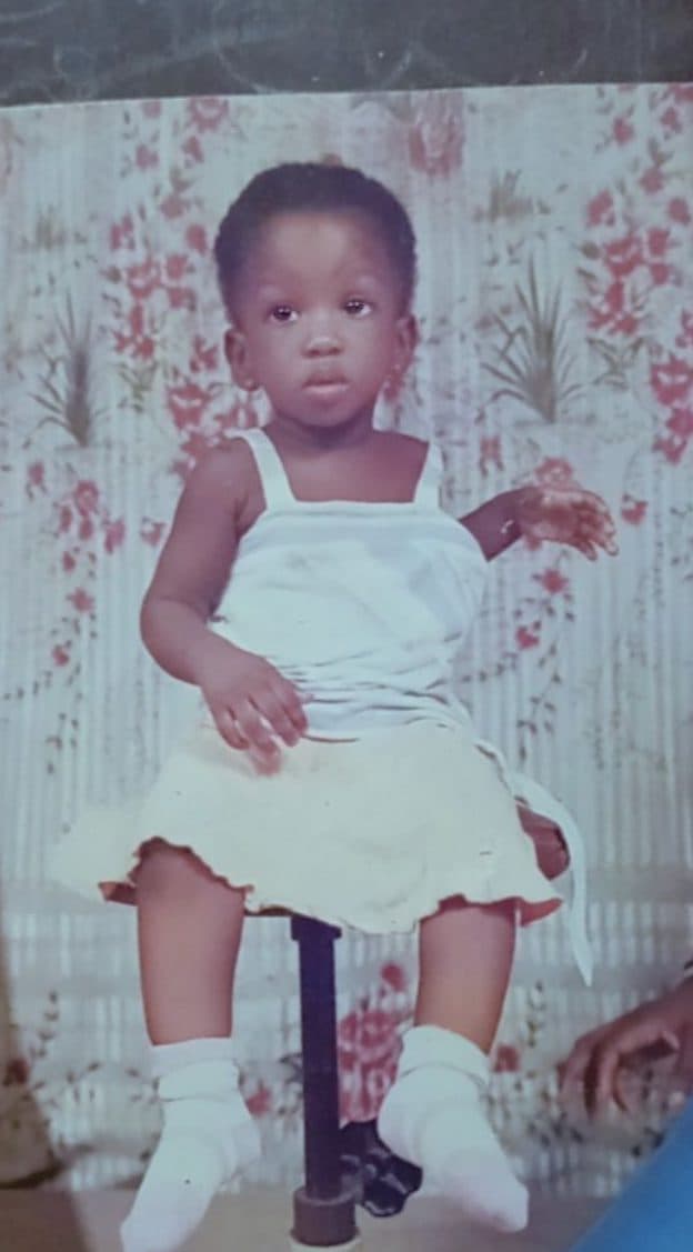 Photo d’enfance : une petite fille en robe blanche, assise sur un tabouret devant un mur fleuri.