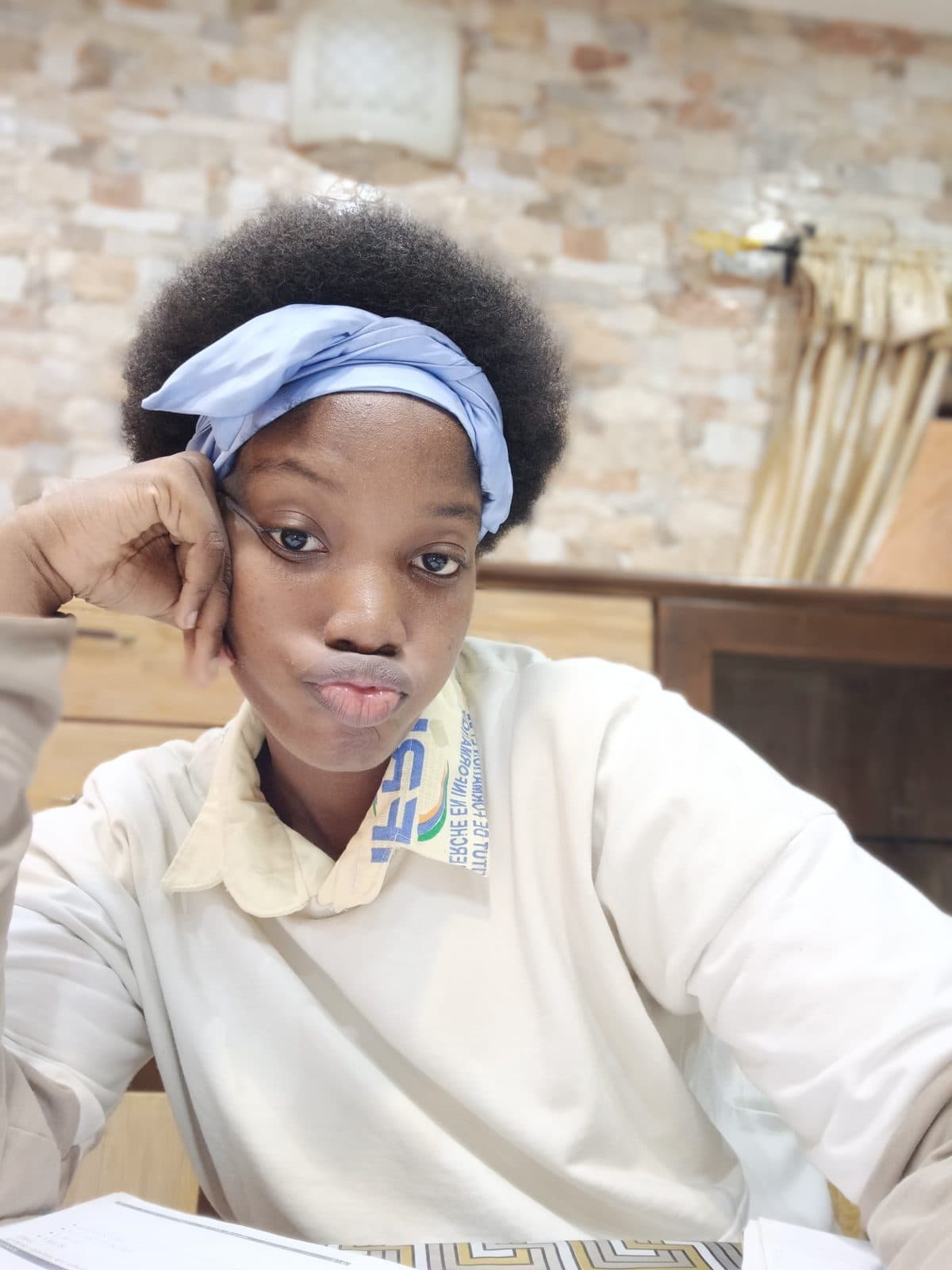Omotola en buste, la joue posée contre la main, un bandeau clair dans les cheveux, devant un mur de briques.