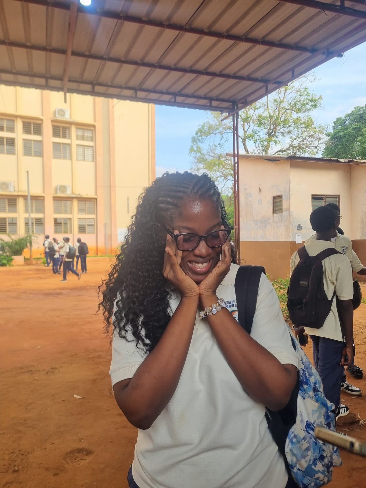 Omotola les mains contre les joues, un grand sourire, devant les bâtiments du campus.